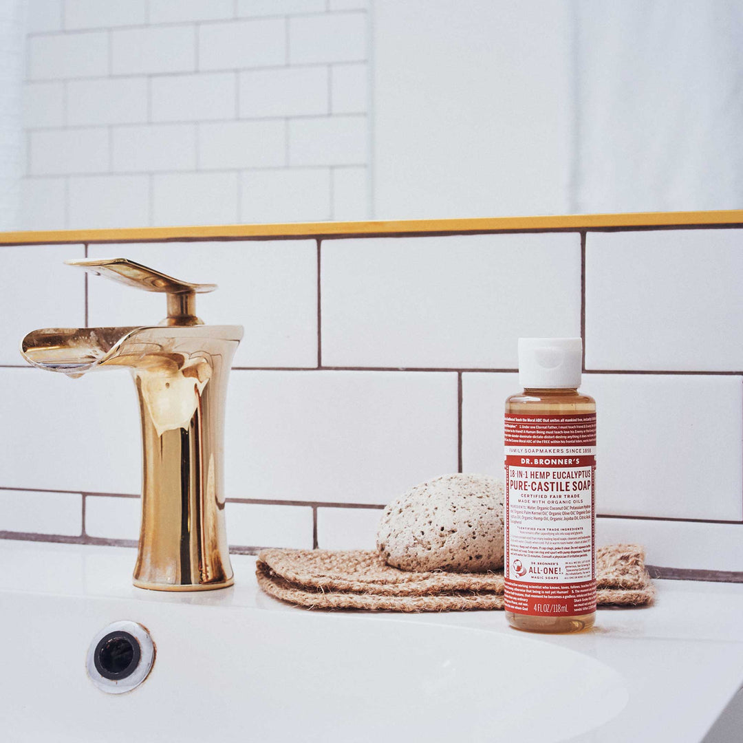 A gold faucet beside a bottle of Dr. Bronners 18 in 1 Hemp Eucalyptus Pure Castile Soap sits on a white bathroom sink with a sponge on a mat [4 oz]