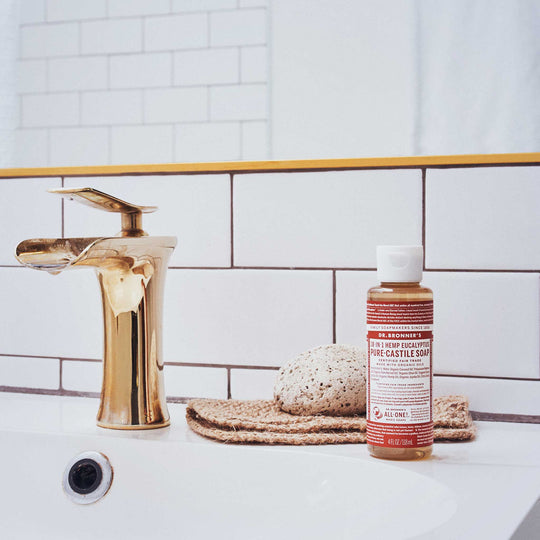A gold faucet beside a bottle of Dr. Bronners 18 in 1 Hemp Eucalyptus Pure Castile Soap sits on a white bathroom sink with a sponge on a mat [4 oz]