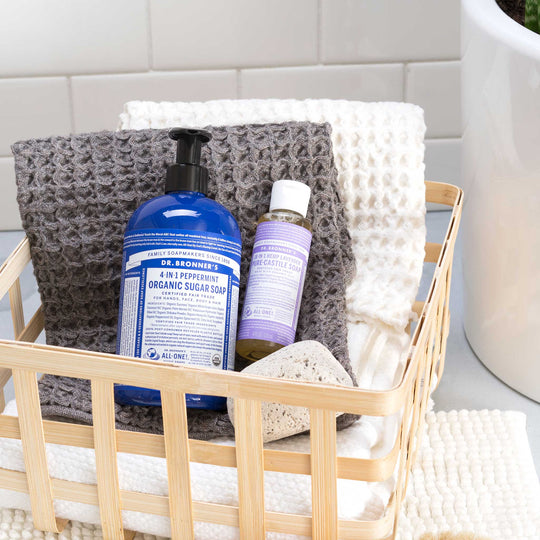 A bamboo basket holds Dr. Bronners 4 in 1 Peppermint Organic Sugar Soap and 18 in 1 Hemp Lavender Pure Castile Soap, atop textured towels beside a white tiled wall. DR. BRONNERS 4 IN 1 PEPPERMINT ORGANIC SUGAR SOAP DR. BRONNERS 18 IN 1 HEMP LAVENDER PURE CASTILE SOAP [4 oz]