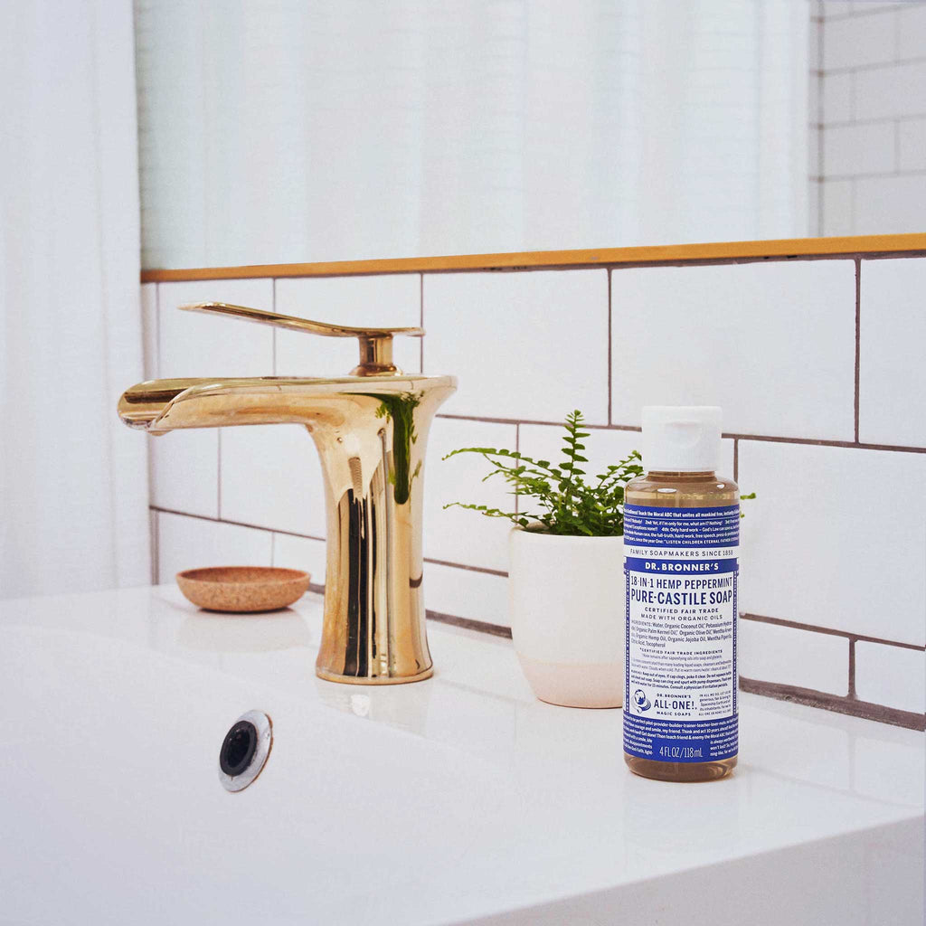 A bottle of Dr. Bronners 18 in 1 Hemp Peppermint Pure Castile Soap sits on a bathroom counter beside a gold faucet and a small potted plant, against tiled walls [4 oz]