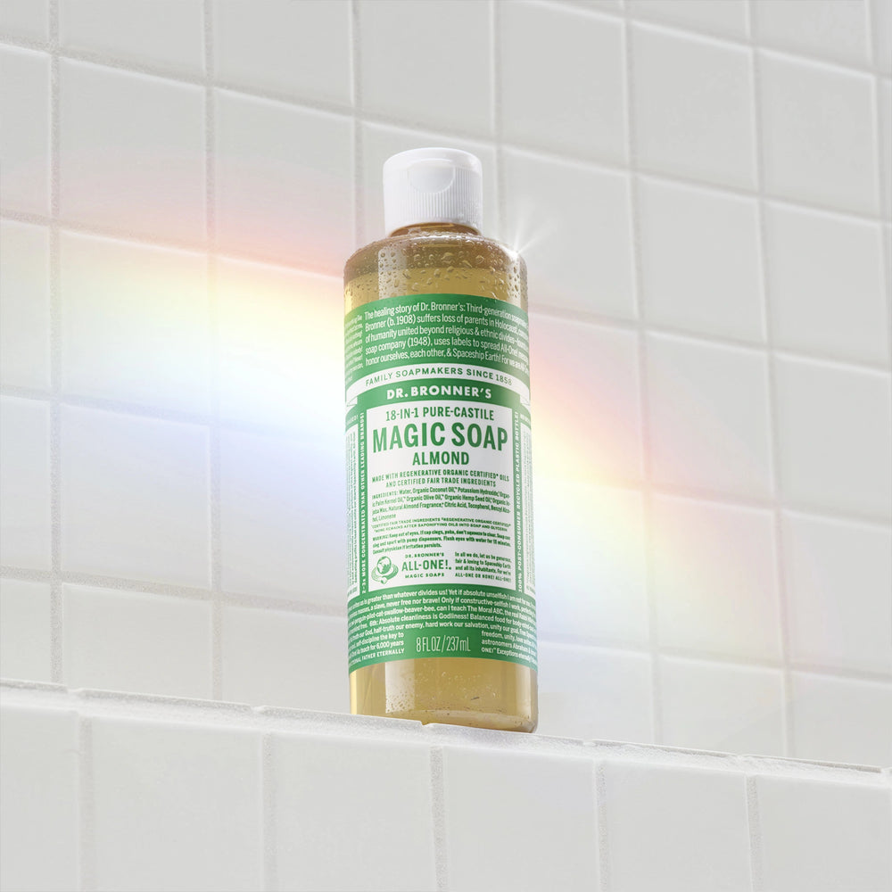 A bottle of Dr. Bronners 18 in 1 Pure Castile Magic Soap stands on a white tiled shelf, illuminated by a rainbow light. Text includes product information and usage instructions [8 oz]