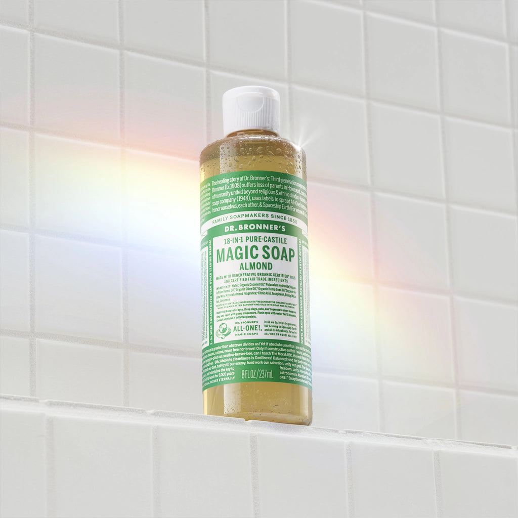 A bottle of Dr. Bronners 18 in 1 Pure Castile Magic Soap stands on a white tiled shelf, illuminated by a rainbow light. Text includes product information and usage instructions [8 oz]