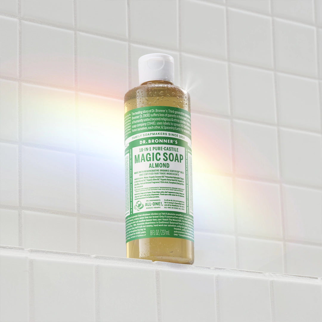 A bottle of Dr. Bronners 18 in 1 Pure Castile Magic Soap stands on a white tiled shelf, illuminated by a rainbow light. Text includes product information and usage instructions [8 oz]