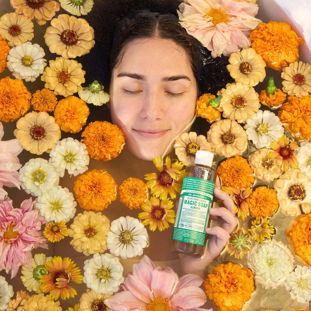 A person lies in a flower filled bath holding a bottle of Dr. Bronners Magic Soap. The colorful flowers create a serene and natural setting for relaxation and skincare. Text on the bottle reads Dr. Bronners 18 in 1 Hemp Peppermint Pure Castile Soap [8 oz]