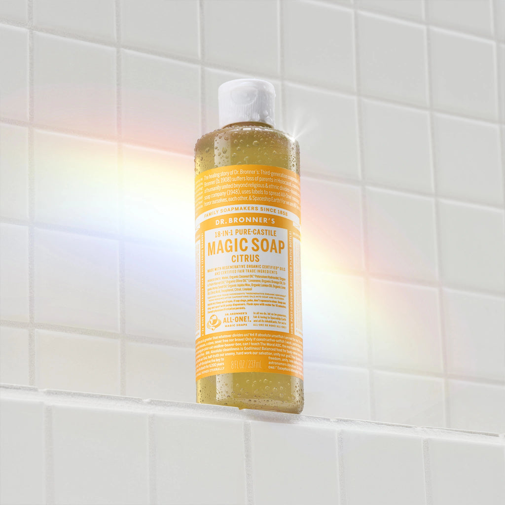 A yellow bottle of Dr. Bronners citrus castile soap sits against a tiled wall, illuminated by a rainbow light reflection. The label features white text detailing the soaps natural ingredients and eco friendly qualities [8 oz]