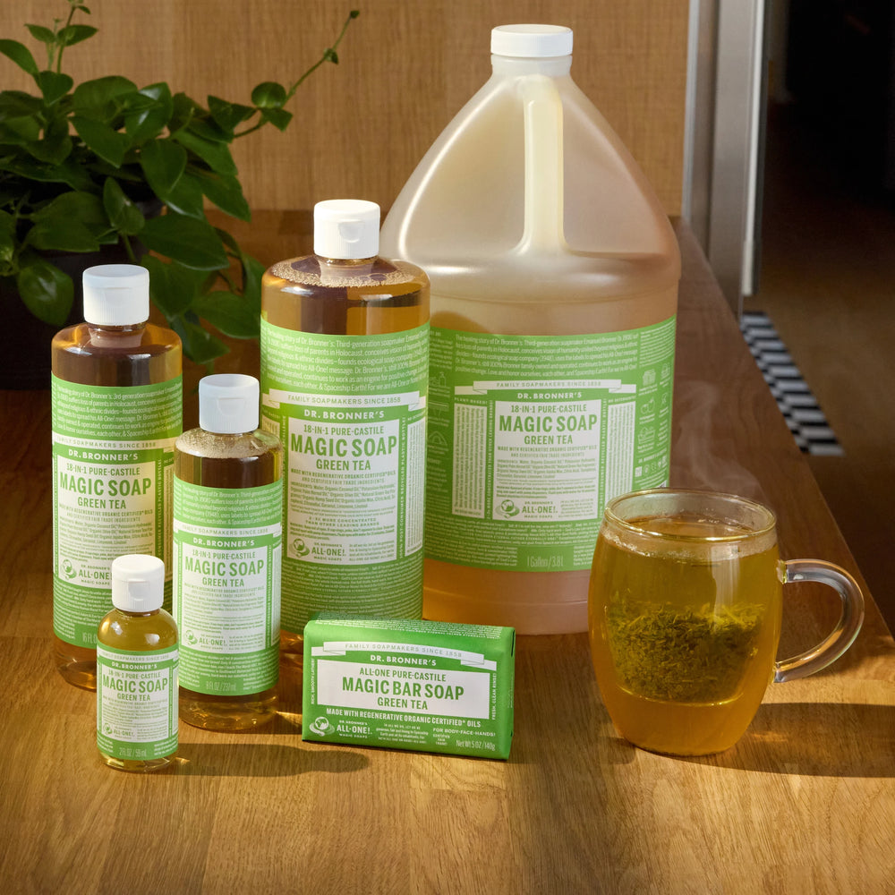 Collection of Dr. Bronner's Magic Soap products on a wooden surface with a glass of liquid. [8 oz, 16 oz, 32 oz, 2 oz, 1 Gallon]