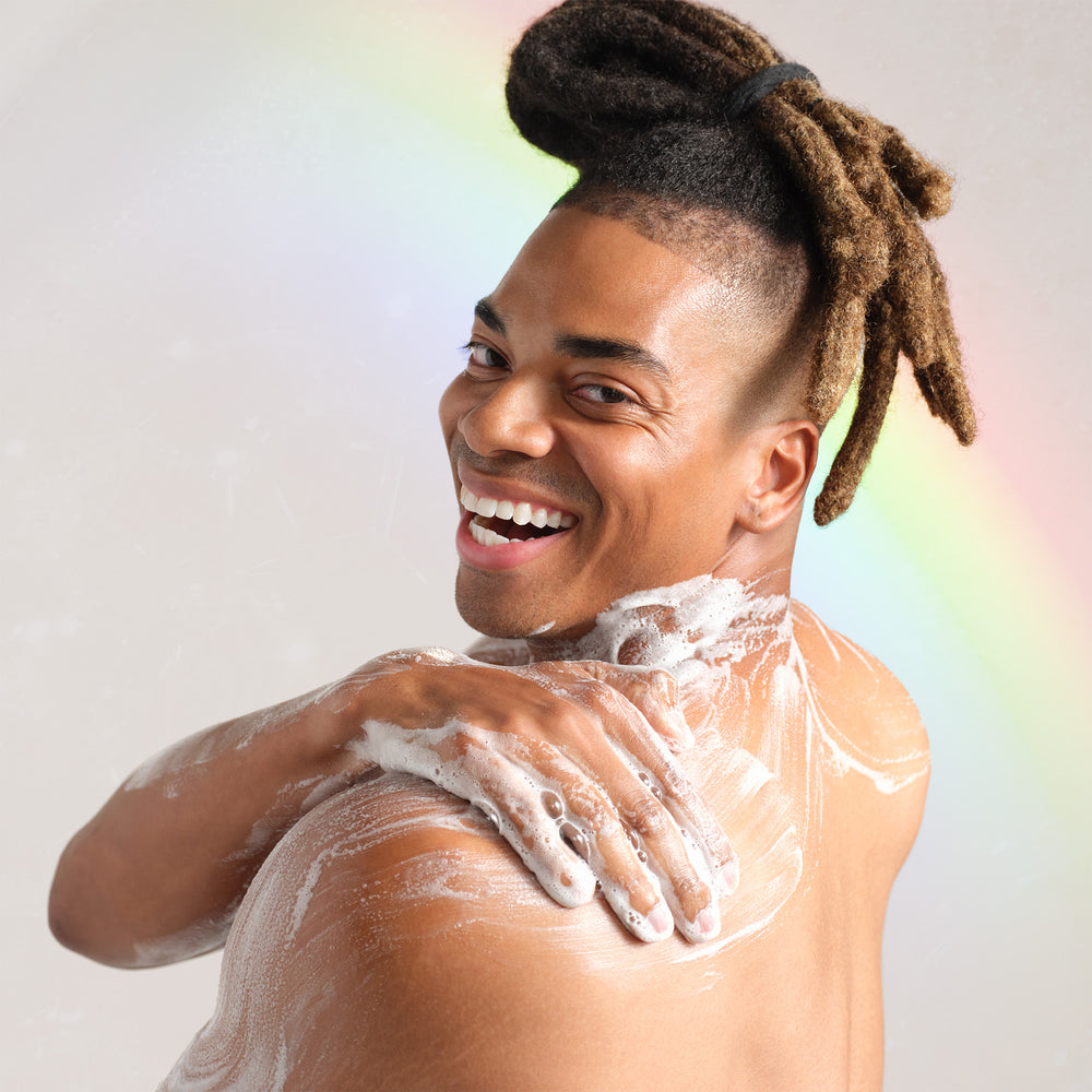A person with a joyful expression applies lathered soap to their shoulder, set against a light background featuring a subtle rainbow. Perfect for illustrating the refreshing use of organic liquid castile soap [2 oz, 3.4 oz, 4 oz, 8 oz, 16 oz, 32 oz, 1 Gallon]