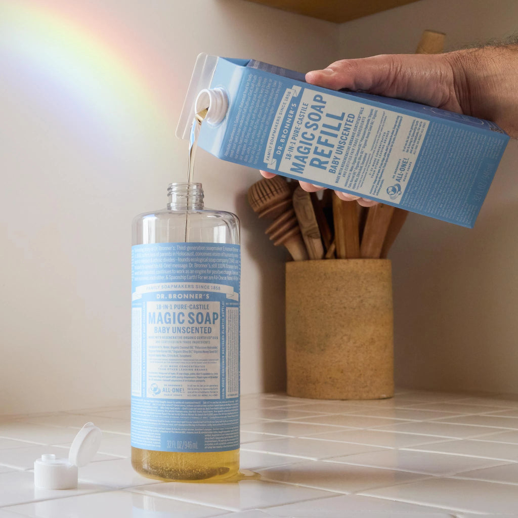A bottle of Dr. Bronners baby unscented castile soap being refilled on a kitchen counter, with a rainbow reflection on the wall. A container with utensils is in the background [32 oz]