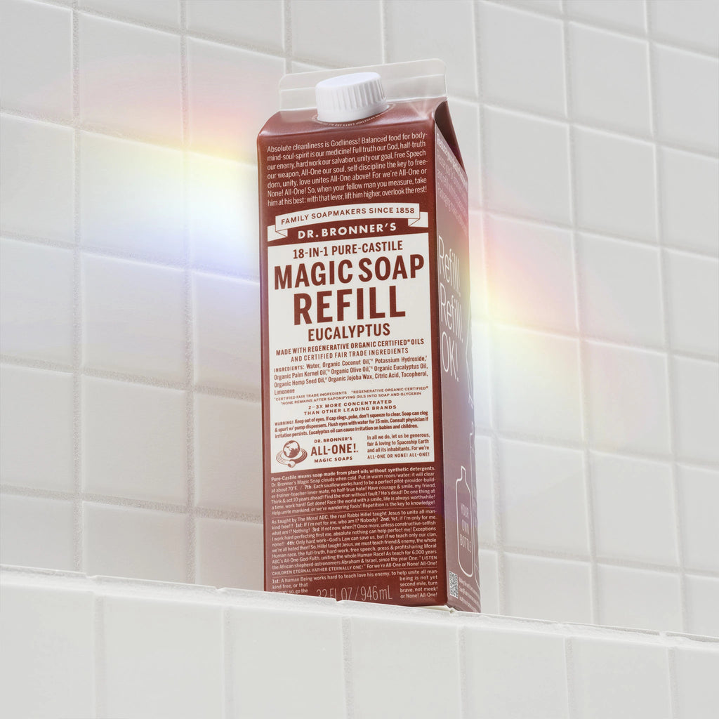 A brown carton labeled MAGIC SOAP REFILL sits on a white tiled shelf, illuminated by a rainbow reflection. The packaging promotes Dr. Bronners natural, eco friendly, organic soap benefits [32 oz]