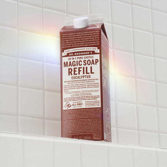 A brown carton labeled MAGIC SOAP REFILL sits on a white tiled shelf, illuminated by a rainbow reflection. The packaging promotes Dr. Bronners natural, eco friendly, organic soap benefits [32 oz]