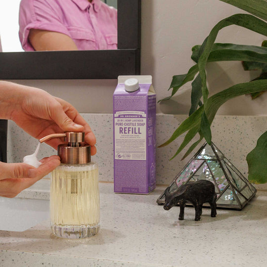 A hand pumps liquid soap from a dispenser on a bathroom counter, flanked by Dr Bronners lavender castile soap carton, a plant, and decorative item. Reflection visible in mirror [32 oz]