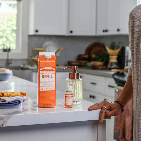 A bottle labeled Dr. Bronners and a liquid container sit on a kitchen countertop. A person stands nearby, with kitchen cabinets and a window in the background [32 oz]