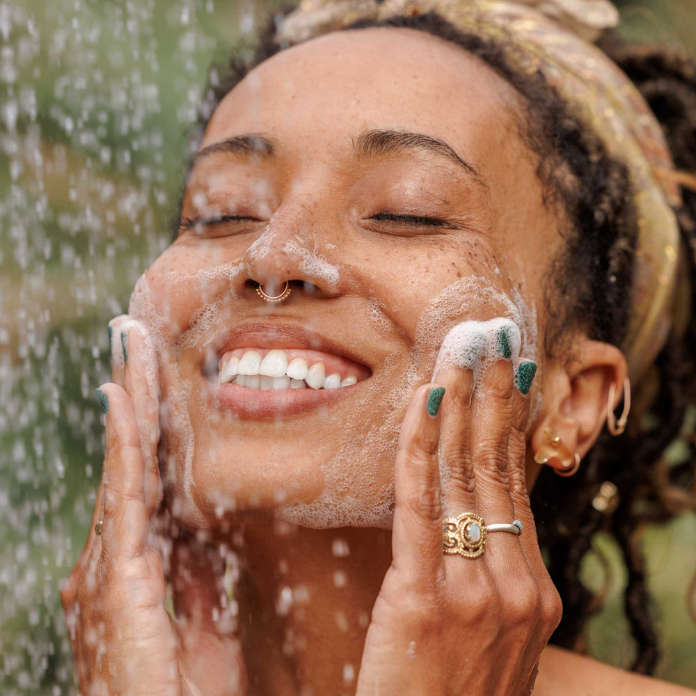 A person gently washes their face, smiling while lathering soap under a shower outdoors, surrounded by greenery. The scene evokes natural skincare, possibly using organic or vegan soap products [2 oz, 3.4 oz, 4 oz, 8 oz, 16 oz, 32 oz, 1 Gallon]