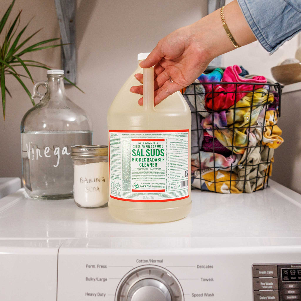 A hand grasps a gallon of Dr. Bronners Sal Suds Biodegradable Cleaner on a laundry machine. Nearby, a bottle labeled Vinegar, a jar of Baking Soda, and a basket of colorful cloths are visible [1 Gallon]