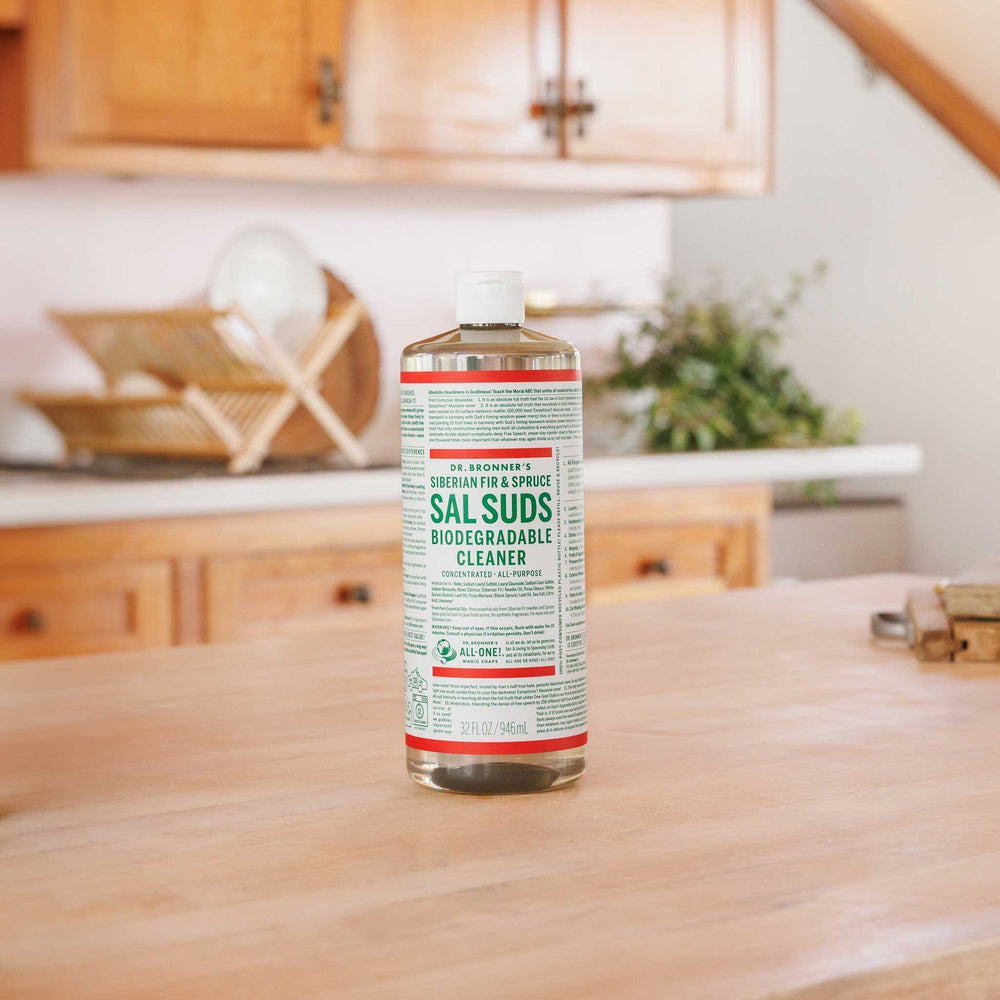 A bottle of Dr. Bronners Sal Suds Biodegradable Cleaner stands on a wooden kitchen counter with cabinets, dishes, and potted plants in the background. Dr. Bronners 16 FL OZ 473 mL [32 oz]