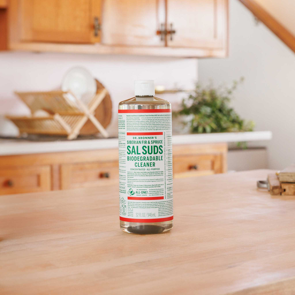 A bottle of Dr. Bronners Sal Suds Biodegradable Cleaner stands on a wooden kitchen counter with cabinets, dishes, and potted plants in the background. Dr. Bronners 16 FL OZ 473 mL [32 oz]