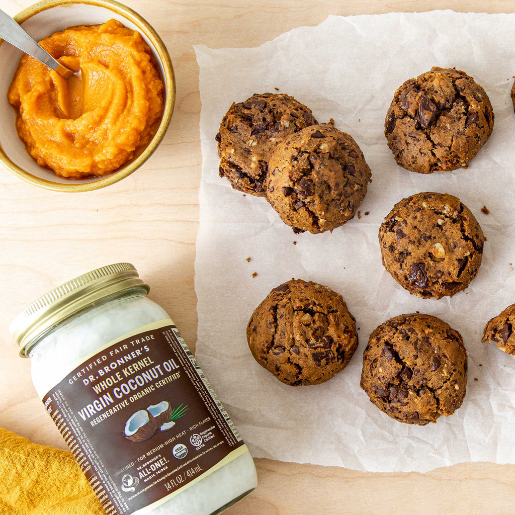 Cookies lie on parchment beside a jar of Dr. Bronners Virgin Coconut Oil. A small bowl of orange puree with a spoon completes the setup on a wooden surface [14 oz]