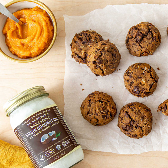 Cookies lie on parchment beside a jar of Dr. Bronners Virgin Coconut Oil. A small bowl of orange puree with a spoon completes the setup on a wooden surface [14 oz]