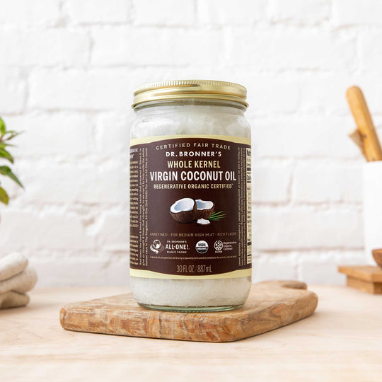 A jar of Dr. Bronners Virgin Coconut Oil, resting on a wooden cutting board in a bright kitchen setting. The label highlights Certified Fair Trade and Regenerative Organic Certified [30 oz]